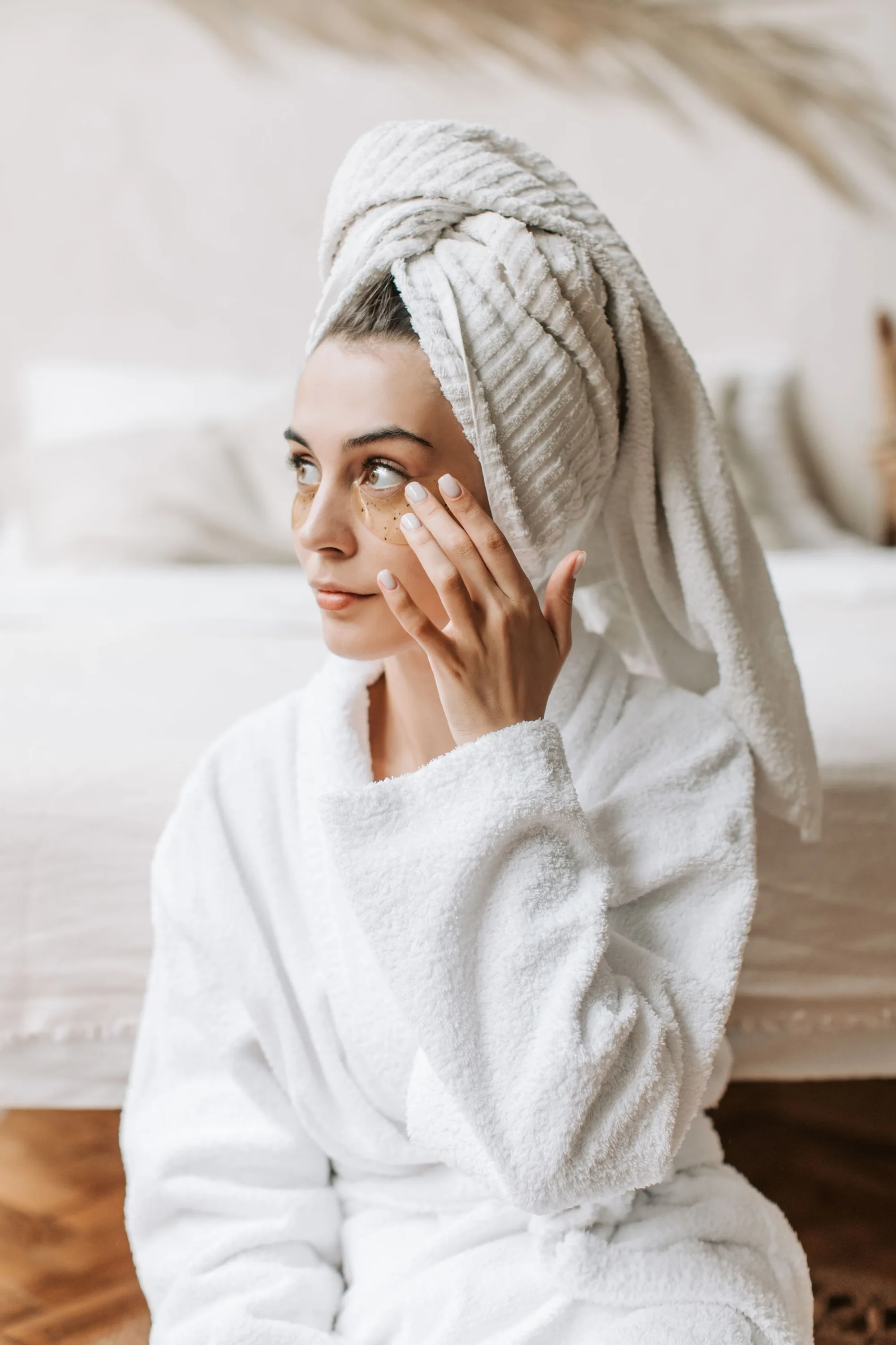 A woman in a bathrobe and towel smoothing cream under her eyes