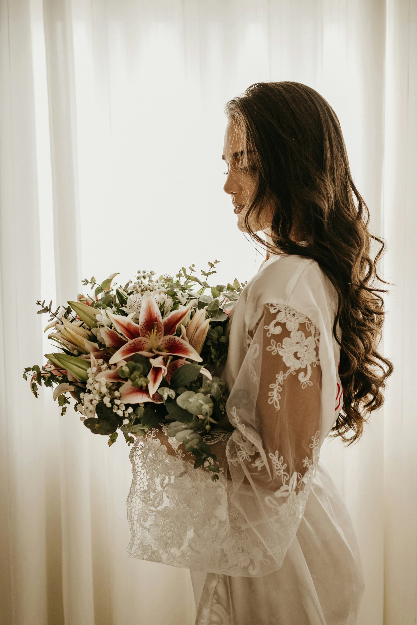 A bride in a lace robe holding her bouquet by a window
