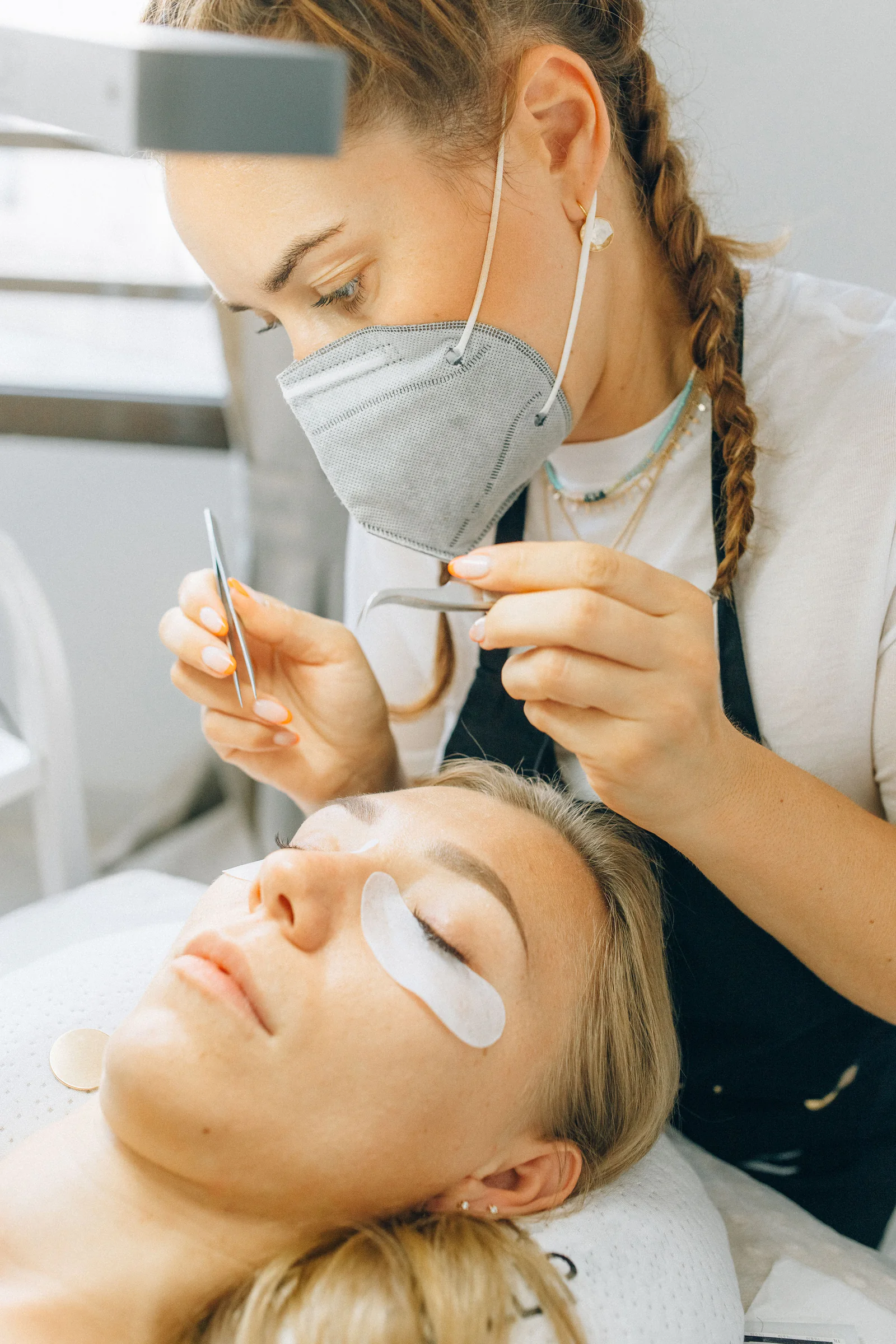 A lash technician working over a client lying on the bed